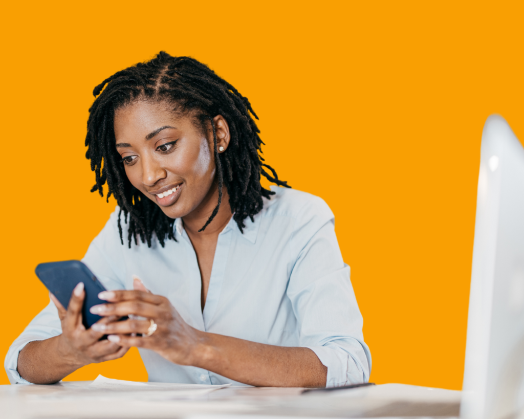 An individual with dreadlocks, wearing a white shirt, is seated at a desk looking at a smartphone with a laptop open in front of them. The background is solid orange.