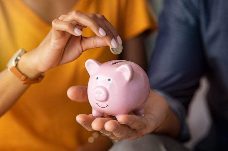“A close-up image of two individuals’ hands, one placing a coin into a pink piggy bank.”