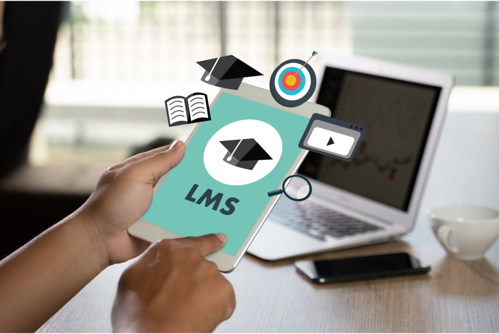A person’s hand holding a smartphone with the screen displaying ‘LMS’ and various education-related icons such as a graduation cap, books, target, and play button floating above it. A laptop and a cup of coffee are in the background on a desk.