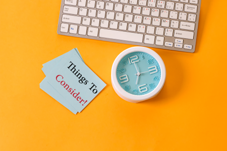 time-management-and-prioritization-concept A white keyboard in the upper left corner on a yellow background, with a blue analog clock showing 10:10 to the right, and a stack of blue cards with “Things To Consider” written in red on the top card.