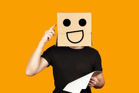 understand-your-needs A person with a cardboard box on their head, which has a drawn face with a smile and eyes, is holding a piece of paper and pointing to their head as if thinking.